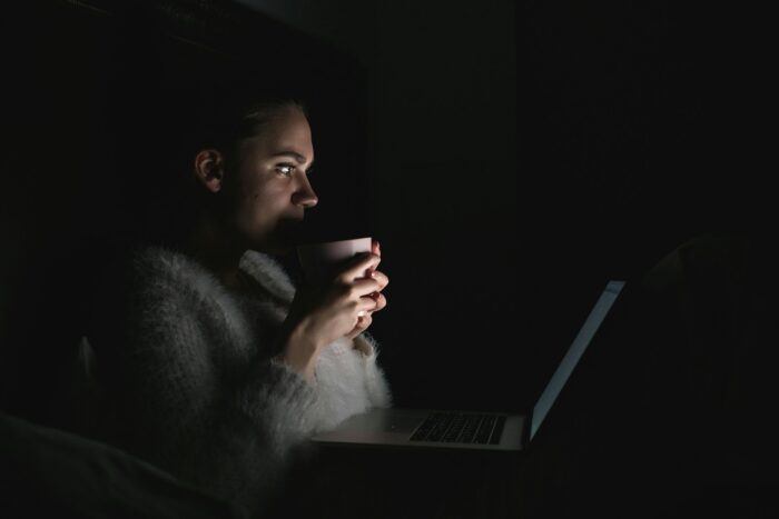 A woman sitting in the dark looking at her laptop screen, holding a cup of coffee.